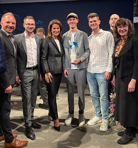 Preisverleihung des TouBy beim Staatsempfang in München - Michaela Kaniber, MdL, überreicht den Jungs von Rhönmomente den Nachwuchspreis 2025 Gruppe von sieben Personen, eine Person hält eine Trophäe. Hintergrund mit dunklem Banner.