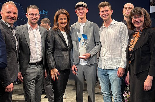 Gruppe von sieben Personen, eine Person hält eine Trophäe. Hintergrund mit dunklem Banner.