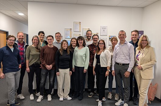Gruppe von 16 Personen steht lächelnd in einem Büroflur vor einer Wand mit Zertifikaten.
