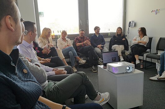 Diskussion beim Treffen der BayTM und dem TVF am 20. Oktober 2025, Nürnberg Eine Gruppe von Menschen sitzt in einem Kreis in einem Besprechungsraum, ein Projektor steht in der Mitte.