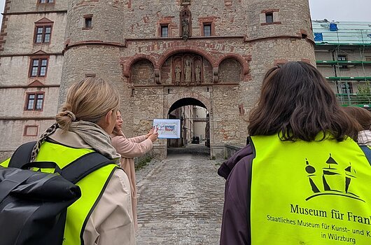 Personen in gelben Westen vor einem historischen Torbogen mit Türmen und Skulpturen.