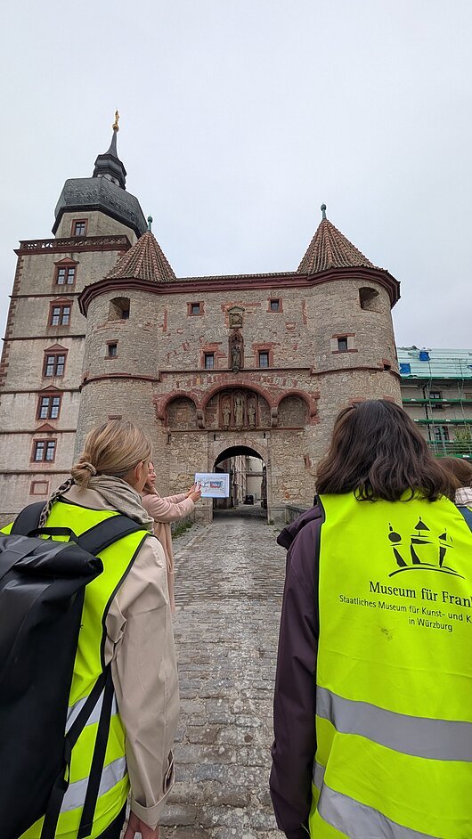 Baustellenbesichtigung für den neuen Standort des Museum für Franken auf der Festung Marienberg, Würzburg Personen in gelben Westen vor einem historischen Torbogen mit Türmen und Skulpturen.