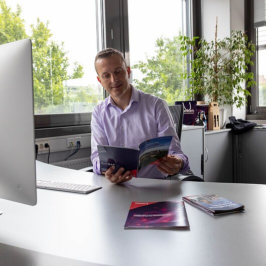 Alexander Hart, Team FrankenTourismus Mann in Hemd sitzt an Schreibtisch und liest Broschüre in hellem Büro mit großen Fenstern.