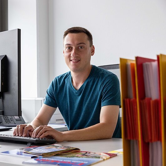 Mann sitzt an Schreibtisch und tippt auf Tastatur vor Monitor in Büro mit Ordnern und Broschüren.
