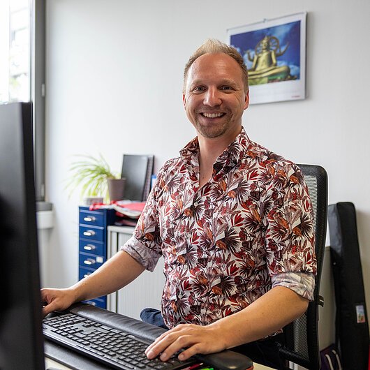 Jörg Hentschel, Team FrankenTourismus Mann mit gemustertem Hemd sitzt lächelnd am Schreibtisch vor Computer in Büro mit Kalender und Pflanze.