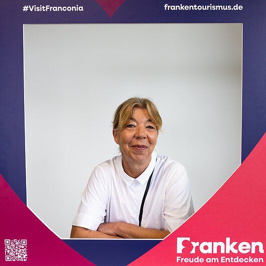 Frau in weißem Hemd hinter blau-rotem Fotorahmen mit Text „Franken Freude am Entdecken“ und Hashtag #VisitFranconia