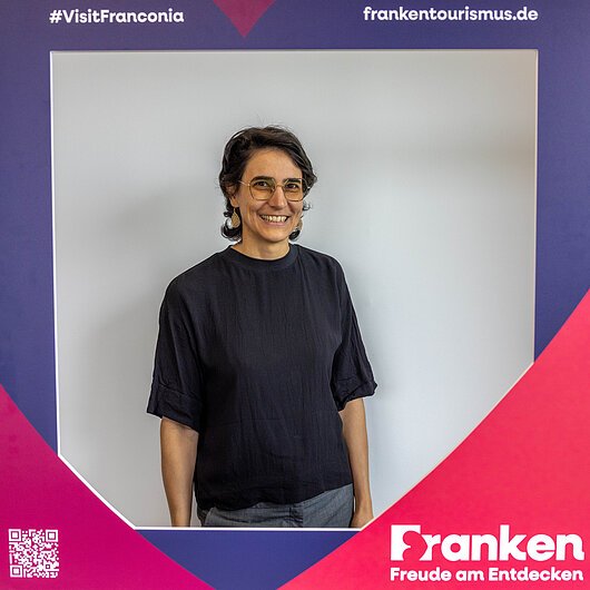 Karolina Klemm, Team FrankenTourismus Frau mit Brille und dunklem Oberteil steht hinter einem Fotorahmen mit Aufschrift „Franken Freude am Entdecken“ und Hashtag #VisitFranconia.