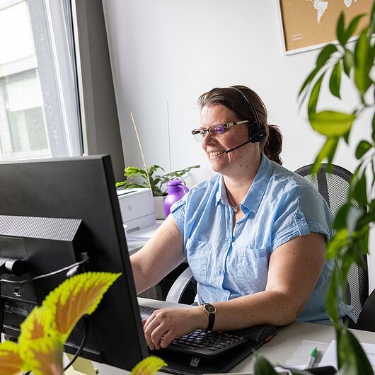 Frau mit Brille und Headset arbeitet am Schreibtisch mit Computer in hellem Büro mit Pflanzen.
