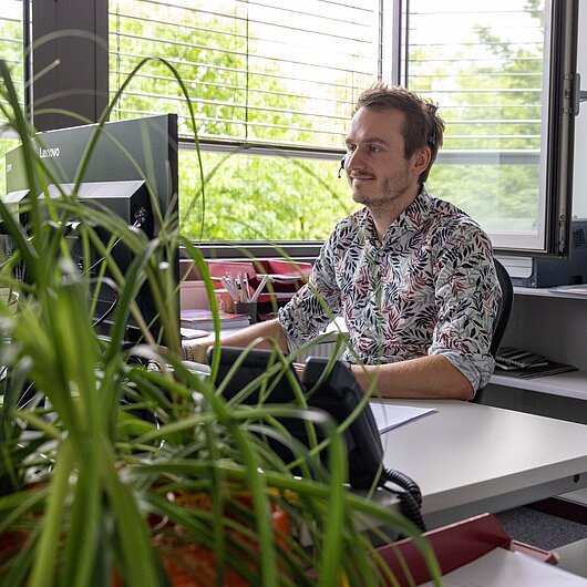 Mann mit Headset sitzt an Schreibtisch vor Computer in Büro mit Pflanzen und offenem Fenster.