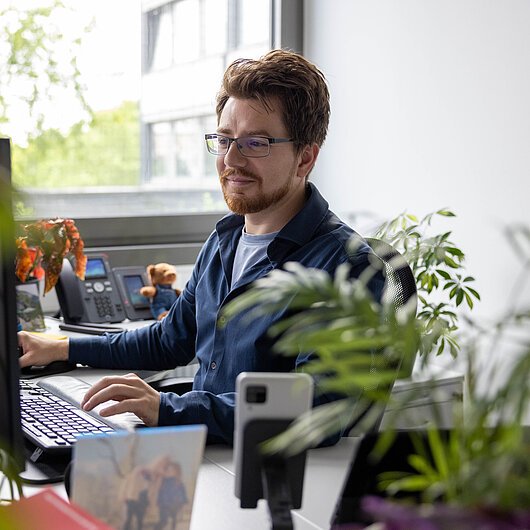 Mann mit Brille arbeitet an einem Computer in einem Büro mit Pflanzen und Telefon im Hintergrund.