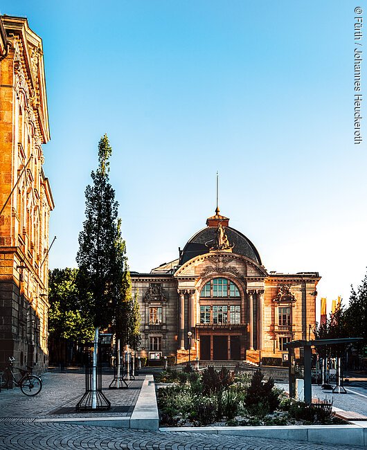 Historisches Gebäude mit Kuppeldach, Bäumen und gepflastertem Platz im Vordergrund bei klarem Himmel