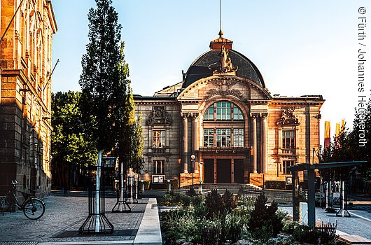 Historisches Gebäude mit Kuppeldach, Bäumen und gepflastertem Platz im Vordergrund bei klarem Himmel