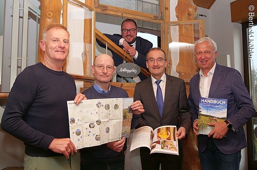 Fünf Männer in Innenraum, vier halten Broschüren, einer lehnt im geöffneten Fensterrahmen mit Schild „Lieblingsplatz“