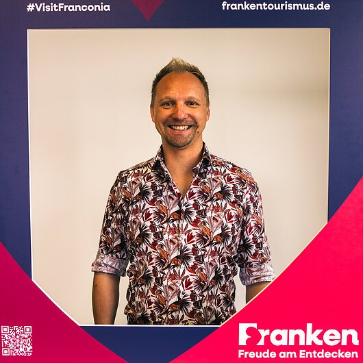 Porträt eines lächelnden Mannes mit kurz geschnittenem Haar und floralem Hemd vor Werberahmen „Franken Freude am Entdecken“.