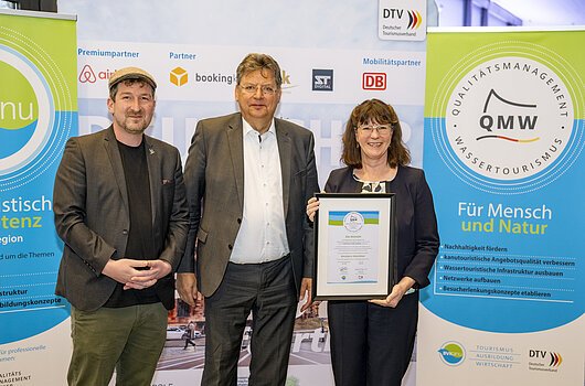 Drei Personen stehen vor Bannern des Qualitätsmanagements Wassertourismus, eine Frau hält eine Urkunde.