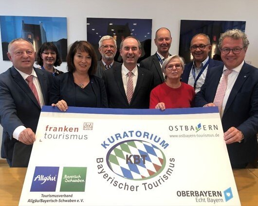 Neun Personen stehen hinter einem großen Banner mit Logos und Schrift zu bayerischem Tourismus.