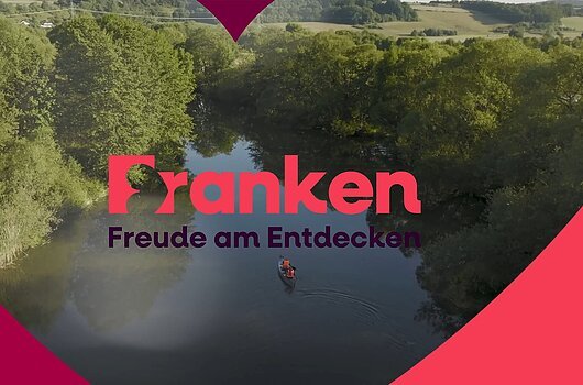 Image-Clip "Reiseland Franken" 2026 Fluss mit zwei Personen im Kanu, umgeben von Bäumen, Text „Franken Freude am Entdecken“ in der Bildmitte.