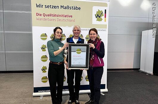 Urkundenübergabe für das Räuberland, Stuttgart (2026) Drei Frauen halten gemeinsam einen gerahmten Zertifikatspreis vor einem Banner der Qualitätsinitiative Wanderbares Deutschland.