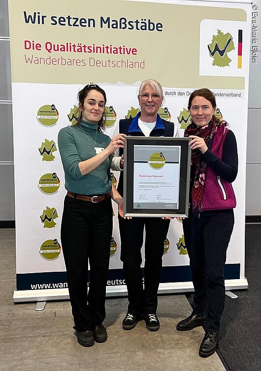Drei Frauen halten gemeinsam einen gerahmten Zertifikatspreis vor einem Banner der Qualitätsinitiative Wanderbares Deutschland.