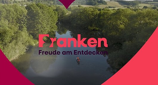 Fluss mit zwei Personen im Kanu, umgeben von Bäumen, Text „Franken Freude am Entdecken“ in der Bildmitte.