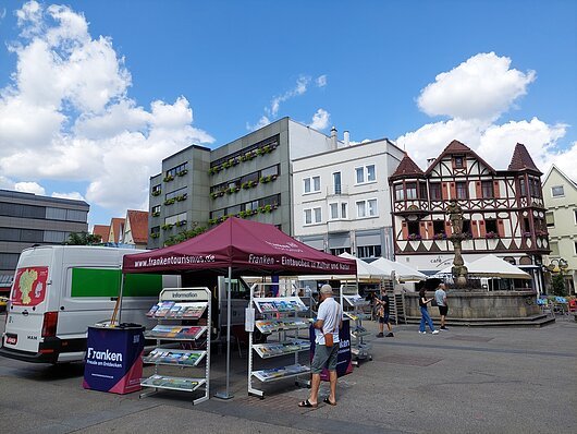 Marktstand mit Informationsmaterial vor modernen und Fachwerkhäusern bei blauem Himmel mit Wolken.