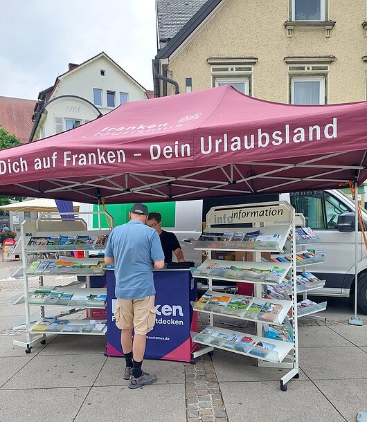 Infostand mit Broschüren unter rotem Zelt mit Aufschrift „Franken – Dein Urlaubsland“ und zwei Personen davor.