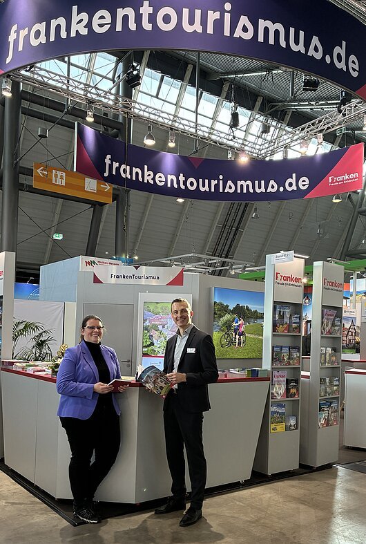Messestand des Tourismusverband Franken mit zwei Personen, Informationsmaterial und Werbebannern in einer Halle