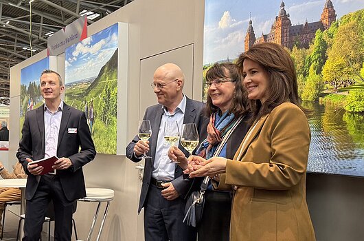 Vier Personen mit Weingläsern stehen auf einer Messe vor Bildern von Landschaften und einem Schloss.