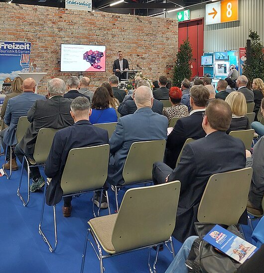 Vortrag von Alexander Hart zur Arbeit des Tourismusverbandes Franken bei der Eröffnung der Freizeit-Messe Nürnberg 2026 Publikum sitzt auf Stühlen vor einem Redner auf einer Messebühne mit Backsteinwand und Präsentationsbildschirm.