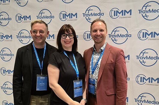 IMM 2026 in Berlin Drei Personen mit Namensschildern vor einer TravMedia- und IMM-Werbewand stehend, lächelnd in die Kamera.