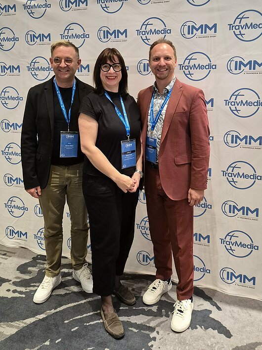 IMM 2026 in Berlin Drei Personen mit Namensschildern vor einer TravMedia- und IMM-Werbewand stehend, lächelnd in die Kamera.