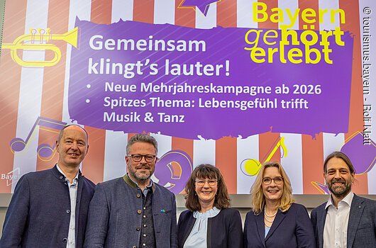 Gruppenbild bei der Pressekonferenz zu "Bayern geHÖRT erlebt" im Rahmen der ITB 2026, Berlin Fünf Personen stehen vor einer Leinwand mit Text zur Mehrjahreskampagne Musik und Tanz in Bayern ab 2026.