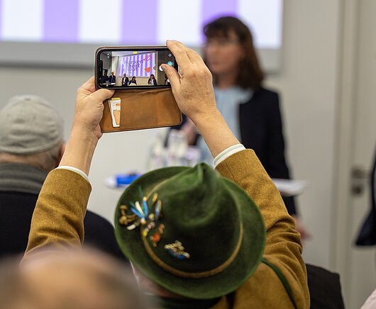 Person mit grünem Hut filmt Podiumsdiskussion mit Smartphone in braunem Lederetui.