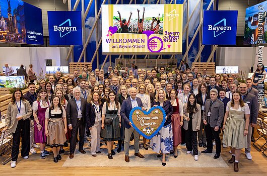 Gruppenfoto von etwa 80 Personen vor Bayern-Messestand mit Schild „Servus aus Bayern“ im Vordergrund.