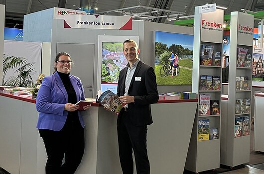 Messestand des Tourismusverband Franken mit zwei Personen, Informationsmaterial und Werbebannern in einer Halle