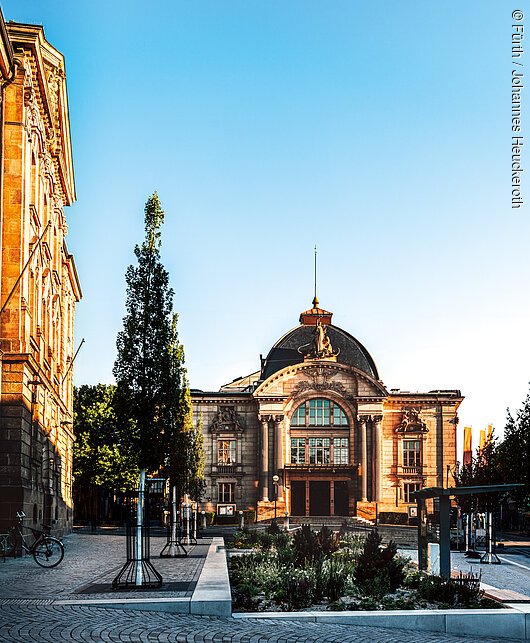 Historisches Gebäude mit Kuppeldach, Bäumen und gepflastertem Platz im Vordergrund bei klarem Himmel