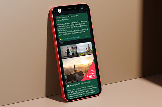 Neuer WhatsApp-Kanal für frankenTourismus Smartphone mit rotem Rahmen zeigt WhatsApp-Chat mit Willkommensnachricht von FrankenTourismus und Bildern aus Franken.