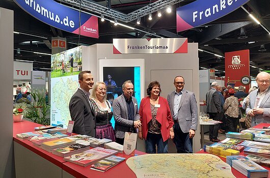 Messerundgang mit Staatsminister Thorsten Glauber bei der Freizeit-Messe 2026 in Nürnberg Fünf Personen stehen an einem Messestand mit Broschüren und Karten, darüber Banner mit der Aufschrift FrankenTourismus.
