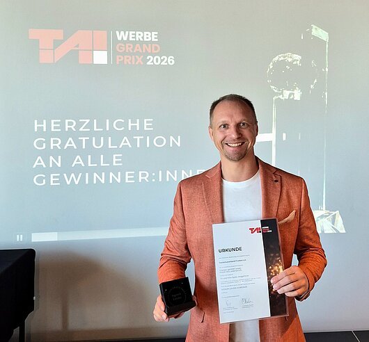 Preisverleihung des T.A.I. Werbe Grand Prix am 28. Mai 2026 (Wien) Mann in orangefarbenem Sakko hält Urkunde und Trophäe vor Projektionswand mit Text zum TAI Werbe Grand Prix 2026.