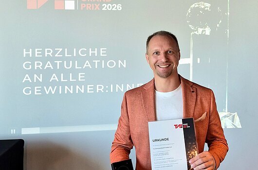 Preisverleihung des T.A.I. Werbe Grand Prix am 28. Mai 2026 (Wien) Mann in orangefarbenem Sakko hält Urkunde und Trophäe vor Projektionswand mit Text zum TAI Werbe Grand Prix 2026.