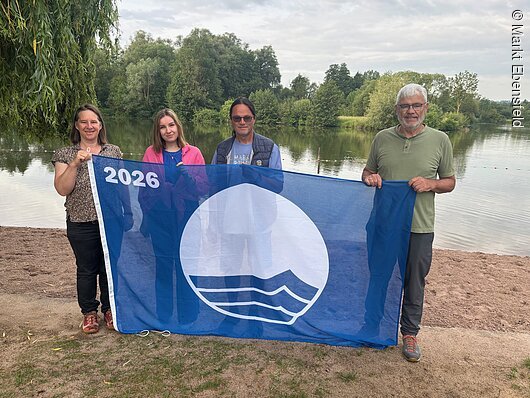Hissen gemeinsam die Blaue Flagge (v.l.): Anne Schmitt (Flussparadies Franken), Lisa Zillig (Verwaltung), Günther Schuberth (Platzwart) und Erster Bürgermeister Bernhard Storath, Markt Ebensfeld Vier Personen stehen an einem Seeufer und halten eine blaue Flagge mit weißem Logo und Jahreszahl 2026.