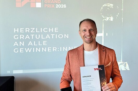 Preisverleihung des T.A.I. Werbe Grand Prix am 28. Mai 2026 (Wien) Mann in orangefarbenem Sakko hält Urkunde und Trophäe vor Projektionswand mit Text zum TAI Werbe Grand Prix 2026.