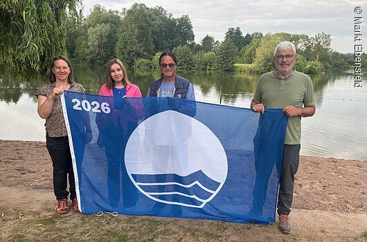 Hissen gemeinsam die Blaue Flagge (v.l.): Anne Schmitt (Flussparadies Franken), Lisa Zillig (Verwaltung), Günther Schuberth (Platzwart) und Erster Bürgermeister Bernhard Storath, Markt Ebensfeld Vier Personen stehen an einem Seeufer und halten eine blaue Flagge mit weißem Logo und Jahreszahl 2026.