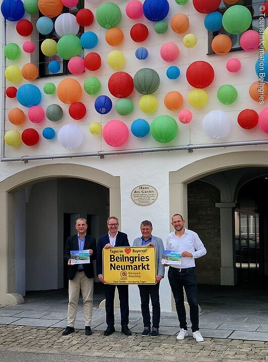 Oberbürgermeister Markus Ochsenkühn (Neumarkt i.d.OPf.) und 1. Bürgermeister Helmut Schloderer (Beilngries) stellten die gemeinsame Gästekarte sowie die Kooperation zwischen im Tagungsbereich Beilngries und Neumarkt i.d.OPf. vor. Vier Männer stehen vor einem weißen Gebäude mit bunten Lampions und halten ein gelbes Schild mit Ortsnamen.