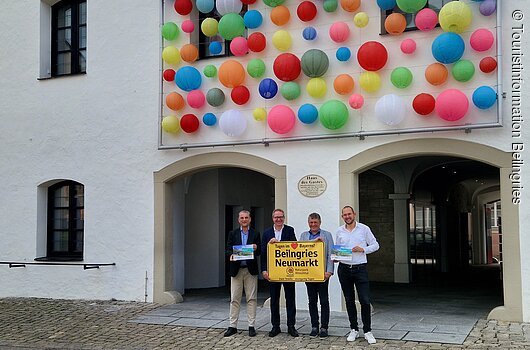 Oberbürgermeister Markus Ochsenkühn (Neumarkt i.d.OPf.) und 1. Bürgermeister Helmut Schloderer (Beilngries) stellten die gemeinsame Gästekarte sowie die Kooperation zwischen im Tagungsbereich Beilngries und Neumarkt i.d.OPf. vor. Vier Männer stehen vor einem weißen Gebäude mit bunten Lampions und halten ein gelbes Schild mit Ortsnamen.