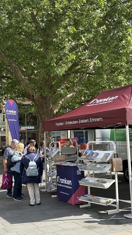 FrankenTour 2026, Wiesbaden Informationsstand mit Broschüren und Banner zur Region Franken unter einem Baum auf einem Platz mit mehreren Personen.