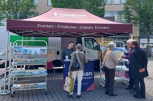 FrankenTour 2026, Darmstadt Infostand mit rotem Zelt und Broschüren vor einem Gebäude, mehrere Personen betrachten Karten und sprechen miteinander.