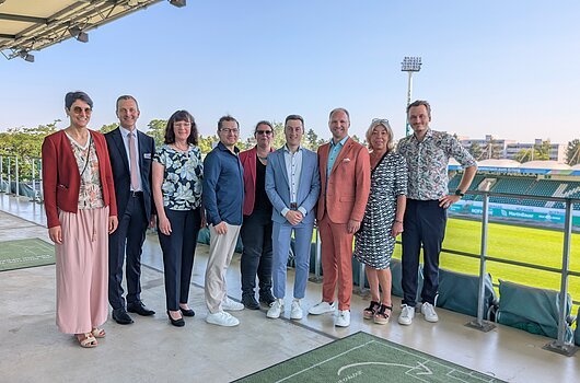 Neun Personen stehen nebeneinander auf einer Stadiontribüne mit Blick auf ein Fußballfeld.