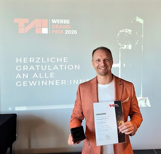 Mann in orangefarbenem Sakko hält Urkunde und Trophäe vor Projektionswand mit Text zum TAI Werbe Grand Prix 2026.
