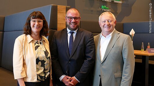 Drei Personen in Businesskleidung stehen nebeneinander und lächeln in einem modernen Raum mit Sitzbank und Tisch.